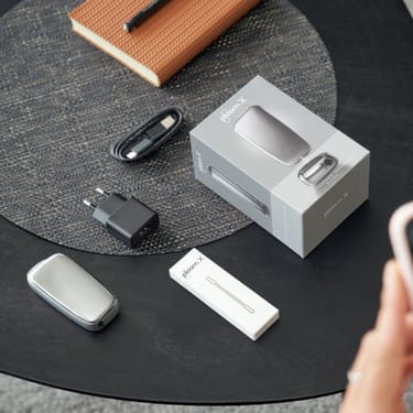 Ploom příslušenství - zařízení Ploom, balení, čistící tyčinka, nabíjecí kabel a adaptér na černošedém pozadí